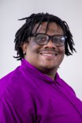 Timotheus (TJ) Gordon Jr. a young black man with long hair. He is smiling and wearing glasses. He is wearing a purple long sleeve shirt

